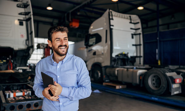 man smiling in a truck workshop holding a tablet surrounded by large commercial trucks