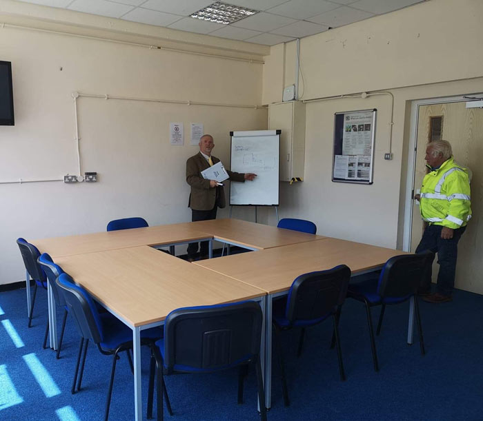 man presenting in a meeting room with a whiteboard and a table with blue chairs